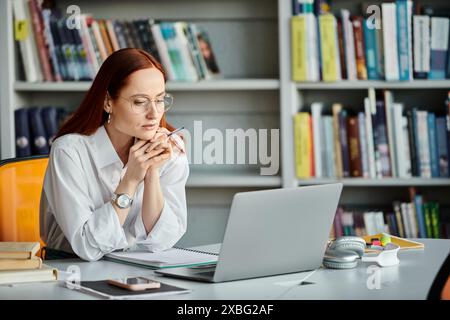 Una tutor rossa sta insegnando una lezione online usando un portatile alla scrivania. Foto Stock