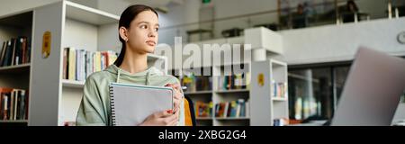 Una ragazza adolescente si trova di fronte a una libreria in una biblioteca, immersa nello studio e nel fare i compiti sul suo portatile dopo la scuola. Foto Stock