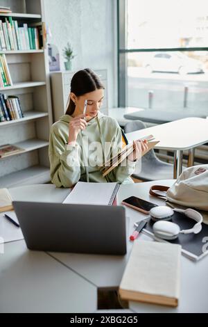 Una ragazza adolescente profondamente pensata, che lavora in multitasking tra il suo notebook e un libro sulla scrivania mentre fa i compiti dopo la scuola. Foto Stock