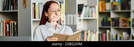 Una donna rossa infilata in un libro, circondata da alti scaffali in una biblioteca, assorbe la conoscenza. Foto Stock