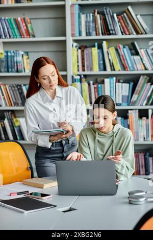 Il tutor di Redhead e la ragazza adolescente si dedicano all'educazione moderna, discutendo delle lezioni scolastiche su un portatile in un ambiente di biblioteca. Foto Stock