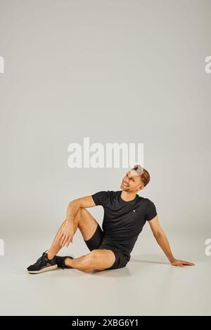 Un giovane atletico siede sul pavimento con una t-shirt nera, persa nel pensiero, in uno studio con sfondo grigio. Foto Stock