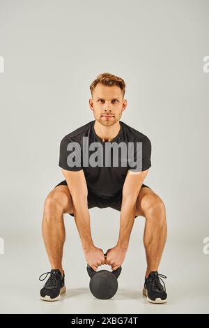 Un giovane sportivo con abbigliamento attivo si accosta con un kettlebell in uno studio con sfondo grigio. Foto Stock