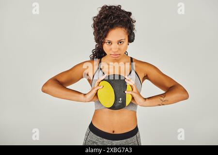 La sportiva riccia afro-americana in abbigliamento attivo si svolge in studio con sfondo grigio, tenendo in mano una palla gialla e nera Foto Stock