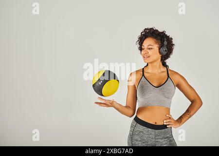 La sportiva riccia afroamericana in abbigliamento attivo mostra forza ed equilibrio mentre tiene in mano una palla gialla e nera vibrante. Foto Stock