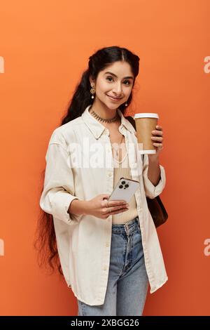 Una giovane ed elegante donna indiana regge felicemente una tazza di caffè su sfondo arancione. Foto Stock