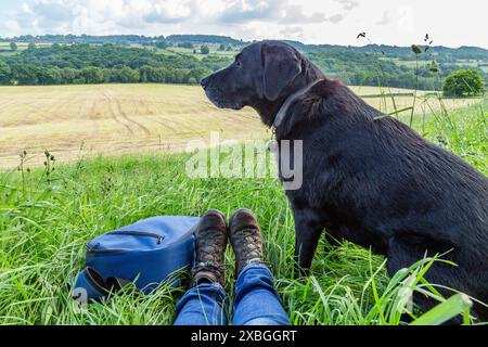 Un labrador retriever nero seduto accanto al suo proprietario ai margini di un campo. Gli stivali da passeggio del proprietario e uno zaino sono accanto al cane. Foto Stock