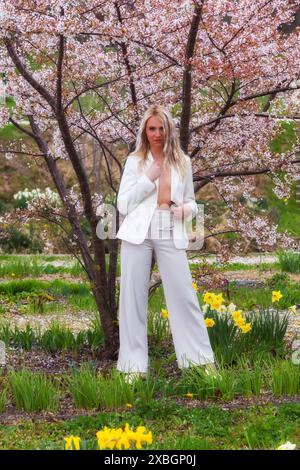 Una giovane donna bionda indossa elegantemente un pantalone bianco con una giacca aperta che mostra il suo petto. È circondata da un'abbondanza di fiori, Foto Stock