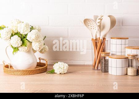 Un set di strumenti per cucinare gli alimenti in un supporto, un set di vasi in ceramica bianca, una caraffa con rami fioriti in un piano di legno. Stile ecologico Foto Stock