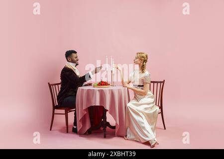 Uomo e donna, vestiti con abiti d'epoca, condividono una cena romantica, bevendo champagne sullo sfondo rosa dello studio. costumi da pranzo del xviii secolo Foto Stock