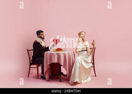 San Valentino Vittoriano. Il gentiluomo condivide il cuore di San Valentino con la signora in un classico abito bianco che guarda al telefono su sfondo rosa. Foto Stock