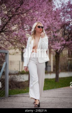 Una modella indossa un costume da pantaloni bianco con una giacca aperta che mostra il suo petto e cammina attraverso un parco pieno di alberi in fiore. Lei indossa il sunglasse Foto Stock