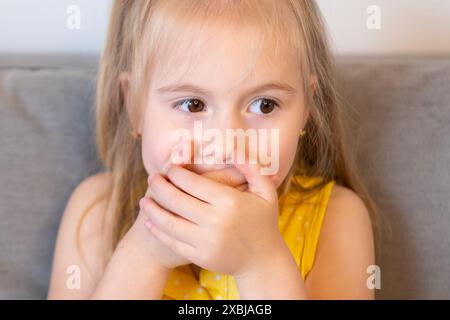 Dolce bambina di 5 anni, timida ragazza bionda, bocca coperta con le mani, lotta per mantenere il segreto e mantenere l'autodisciplina, difficoltà di controllo, mente yo Foto Stock