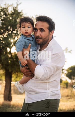 Un bambino di un anno tenuto prigioniero da suo padre con alberi e bokeh sullo sfondo, catturando un momento emozionante nella festa del papà Foto Stock