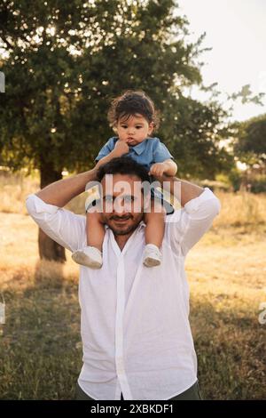 Un bambino di un anno seduto sulle spalle di suo padre con alberi e bokeh sullo sfondo, catturando un momento speciale nella festa del papà Foto Stock