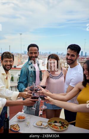 Sorridenti giovani multietnici che brindano bicchieri di vino in piedi per festeggiare i compleanni sul tetto. Foto Stock