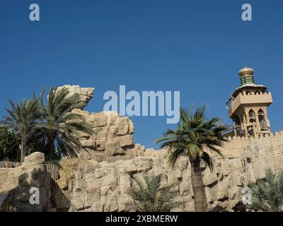 Edifici dal design tradizionale con palme e formazioni rocciose sotto un cielo azzurro, Dubai, Emirati Arabi Foto Stock