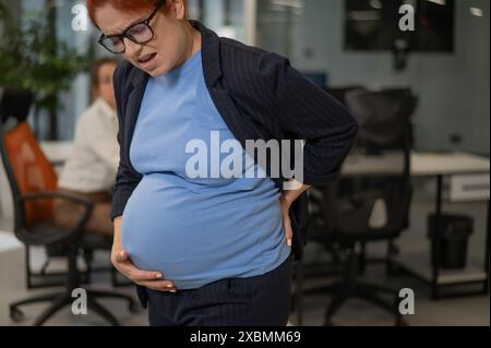 Una donna incinta soffre di dolore e tiene lo stomaco mentre si trova in mezzo all'ufficio. Foto Stock