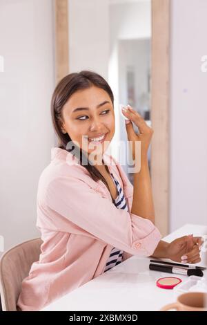 Felice donna birazziale che applica il trucco, sorride mentre guarda il suo riflesso Foto Stock