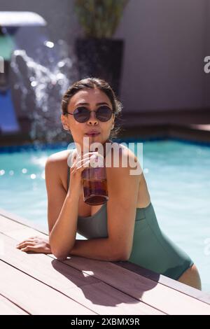 Felice donna birazziale con occhiali da sole che si rilassa a bordo piscina, sorseggiando una bevanda fresca Foto Stock