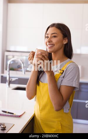 Felice donna birazziale che si gusta un caffè in una cucina moderna con ripiani eleganti Foto Stock