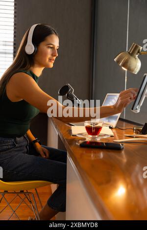 Donna caucasica sorridente con le cuffie che usano il tablet e registrano un podcast Foto Stock