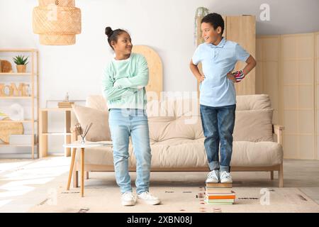 Divertente piccolo ragazzo afroamericano sui libri più alto di sua sorella a casa Foto Stock