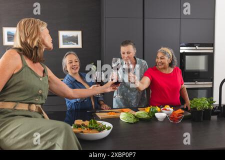 Gruppo di donne anziane che si godono il vino e preparano i pasti insieme in una cucina moderna Foto Stock