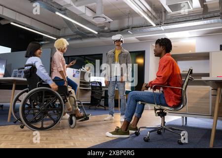 Colleghi d'affari in ufficio che guardano un uomo che usa il visore vr, una donna asiatica in sedia a rotelle, inclusività Foto Stock