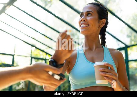 Donna birazziale in abbigliamento sportivo che regge una tazza di caffè pagando con smartwatch Foto Stock