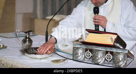 Prete che si prepara per una cerimonia di battesimo, tenendo un microfono e ponendo la mano su una ciotola d'argento piena di acqua Santa Foto Stock