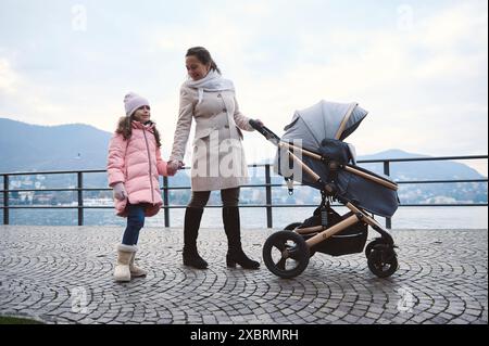 Madre e figlia si godranno una passeggiata con un passeggino lungo una passeggiata panoramica sul lago in una fresca giornata invernale. Attività per famiglie e attività all'aperto nel b Foto Stock