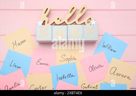 Cubi con la frase Hello Baby e adesivi di carta con nomi diversi su sfondo in legno rosa, piatto. Scegliere il nome del bambino Foto Stock