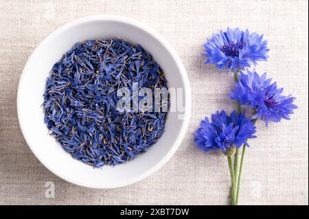 Petali di fiordaliso blu essiccati in una ciotola bianca su tessuto di lino. Fiori commestibili di Centaurea cyanus, noto anche come bottone di scapolo. Foto Stock