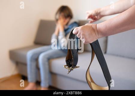 Donna con cintura in pelle in mano affronta il bambino sul divano, disciplina e punizione parentale autoritaria, conflitto familiare e gap generazionale, Pa Foto Stock