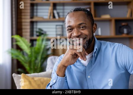 Ritratto di un uomo felice seduto su un divano, con una camicia blu, sorridente e rilassato in un accogliente soggiorno. Foto Stock