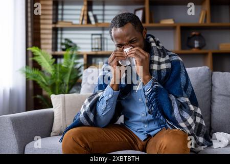 Un uomo starnuto in un tessuto, avvolto in una coperta e seduto su un divano. Descrive la malattia, il raffreddore, l'influenza e il comfort domestico. Foto Stock