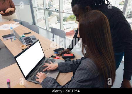 Un uomo e una donna stanno lavorando insieme guardando uno schermo vuoto per laptop in ufficio. Il portatile è aperto sulla scrivania, la donna è seduta a scrivere e il Foto Stock