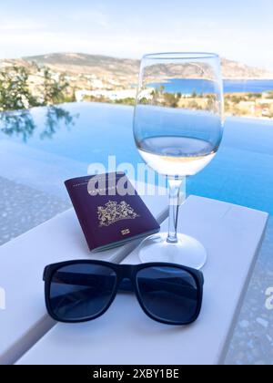 Una pittoresca immagine di viaggio con occhiali da sole, vino bianco e un passaporto su un tavolo, sullo sfondo mozzafiato di un'isola greca e infini Foto Stock