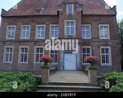 Grande edificio in mattoni a più piani con molte finestre, due vasi di fiori con fiori rossi all'entrata e tempo soleggiato, ochtrup, muensterland Foto Stock