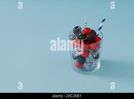 Ciliegie rosse, cubetti di ghiaccio e cannuccia da bere in tazza di vetro su sfondo azzurro. Concetto minimo di cocktail drink. Composizione creativa estiva. Foto Stock