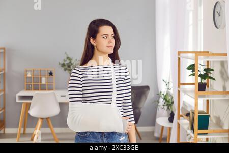 Giovane donna con un infortunio alla mano che indossa un'imbracatura per il braccio in piedi nel soggiorno di casa Foto Stock