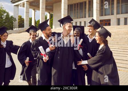 Gruppo di laureati multirazziali felici e risi che abbracciano all'aperto con un diploma. Foto Stock