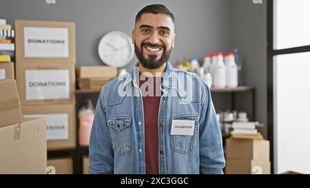 Un uomo sorridente barbuto con un distintivo di "volontario" si trova in un magazzino pieno di scatole per donazioni. Foto Stock