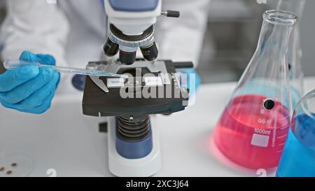 Un ricercatore di guanti utilizza una pipetta su un microscopio accanto a matracci colorate da laboratorio in laboratorio Foto Stock