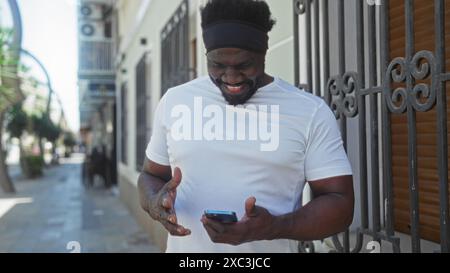 Bel giovane afroamericano con la barba che controlla il telefono su una strada urbana, vestito con una camicia bianca informale, che si gode un ambiente urbano all'aperto Foto Stock
