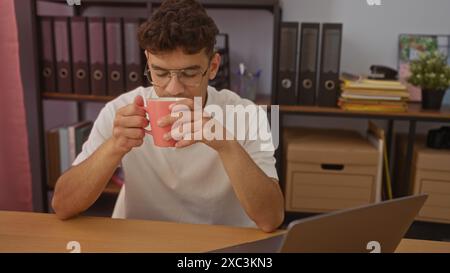 Giovane ispanico che beve caffè in un ufficio, catturato in un momento di riflessione mentre si siede a una scrivania con un portatile, circondato da un raccoglitore organizzato Foto Stock