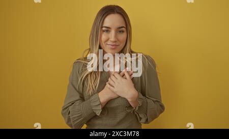 Bella giovane donna bionda in un abito verde oliva con le mani sul petto, isolata su uno sfondo giallo. Foto Stock
