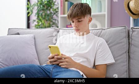 Adolescente biondo con una t-shirt bianca casual e jeans, che utilizza uno smartphone mentre si siede su un divano grigio all'interno. Foto Stock