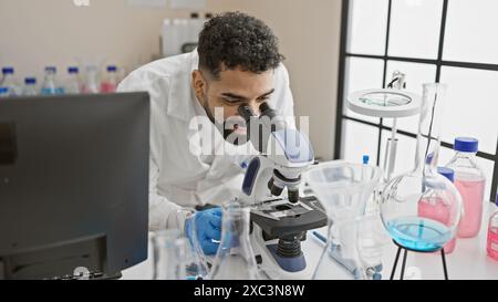 Un giovane ispanico con la barba esamina i campioni utilizzando un microscopio in laboratorio. Foto Stock